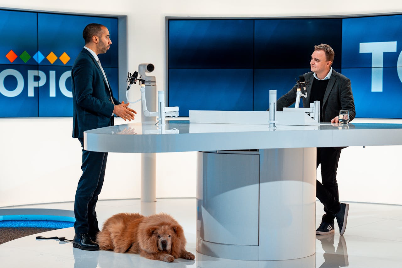 services-03 Two hosts discussing at a modern TV studio with a dog laying by. Engaging professional setup.